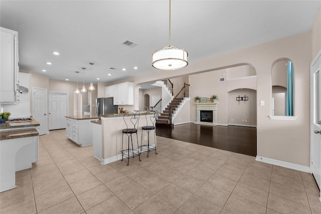 kitchen with light tile patterned flooring, an island with sink, stainless steel fridge, white cabinets, and hanging light fixtures