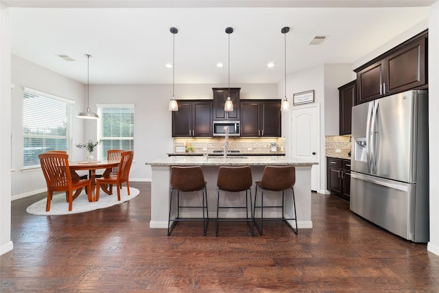 kitchen featuring hanging light fixtures, a kitchen island with sink, light stone counters, stainless steel appliances, and dark wood-type flooring