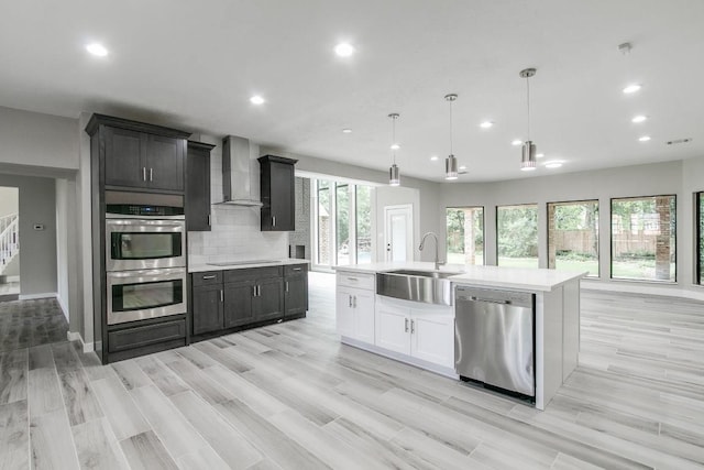 kitchen with sink, wall chimney exhaust hood, stainless steel appliances, an island with sink, and pendant lighting