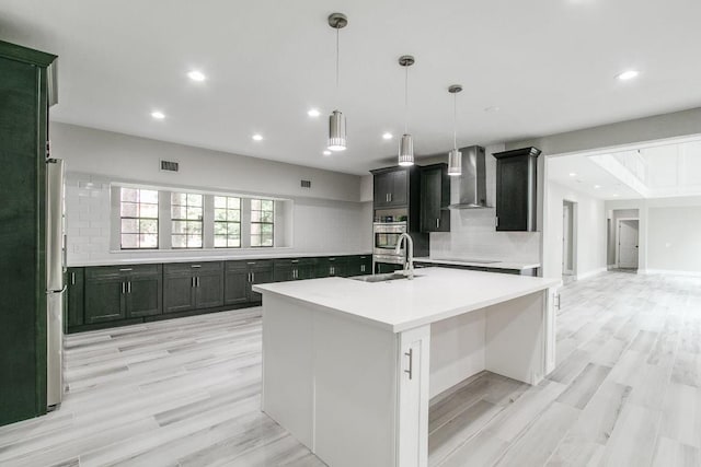 kitchen featuring light wood-type flooring, tasteful backsplash, wall chimney exhaust hood, decorative light fixtures, and a center island with sink