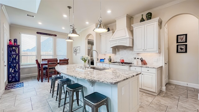 kitchen with premium range hood, light stone counters, sink, decorative light fixtures, and a center island with sink