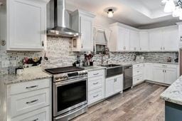 kitchen featuring decorative backsplash, stainless steel appliances, wall chimney range hood, white cabinets, and hardwood / wood-style floors