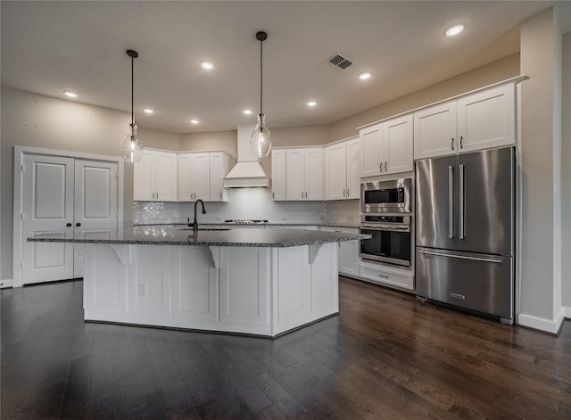 kitchen featuring premium range hood, white cabinetry, an island with sink, stainless steel appliances, and decorative backsplash