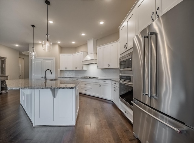 kitchen with custom exhaust hood, white cabinetry, stainless steel appliances, an island with sink, and hanging light fixtures