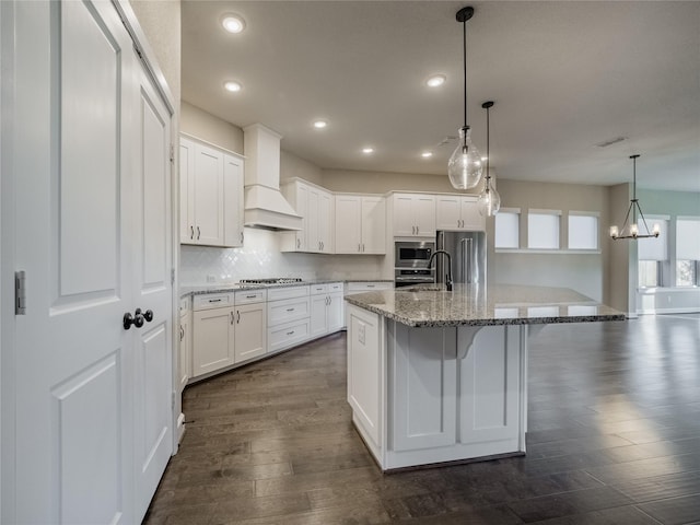 kitchen featuring white cabinets, appliances with stainless steel finishes, custom exhaust hood, a kitchen island with sink, and a chandelier