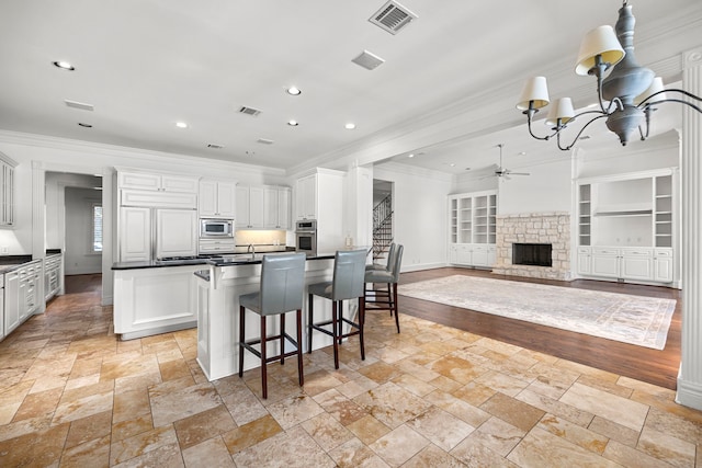 kitchen featuring ceiling fan with notable chandelier, built in appliances, built in features, white cabinetry, and a stone fireplace