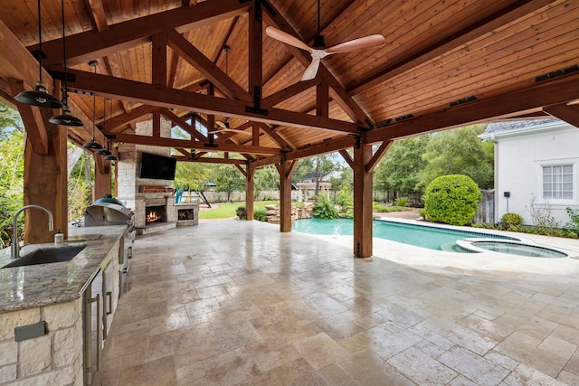 view of swimming pool featuring an outdoor kitchen, an outdoor stone fireplace, ceiling fan, sink, and a gazebo