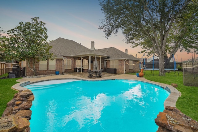 pool at dusk with a trampoline, a patio area, and a lawn