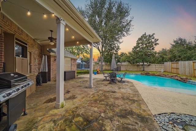 pool at dusk with area for grilling, ceiling fan, a patio area, and a trampoline