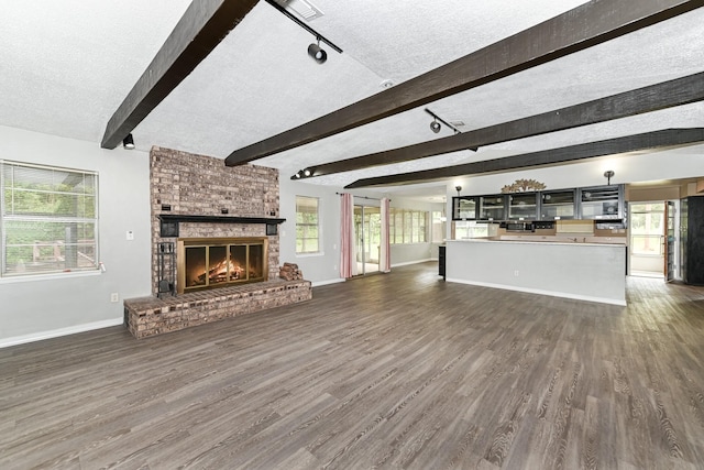 unfurnished living room with dark wood-type flooring, a brick fireplace, a textured ceiling, beamed ceiling, and a healthy amount of sunlight