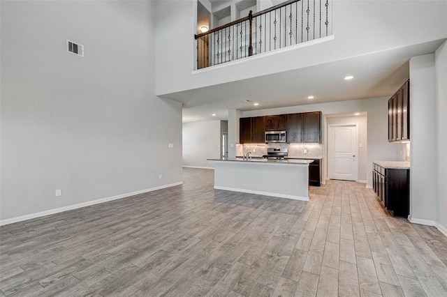 kitchen with appliances with stainless steel finishes, light wood-type flooring, a towering ceiling, dark brown cabinets, and a kitchen island with sink