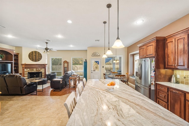 kitchen with backsplash, a stone fireplace, ceiling fan, stainless steel fridge, and decorative light fixtures