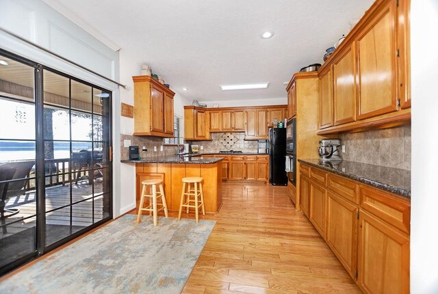 kitchen featuring black refrigerator, dark stone countertops, light wood-type flooring, tasteful backsplash, and a kitchen bar