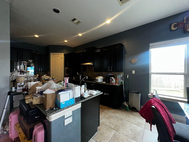 kitchen featuring dark stone countertops, stainless steel microwave, light tile patterned floors, and an island with sink