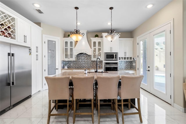 kitchen with backsplash, pendant lighting, built in appliances, white cabinets, and an island with sink