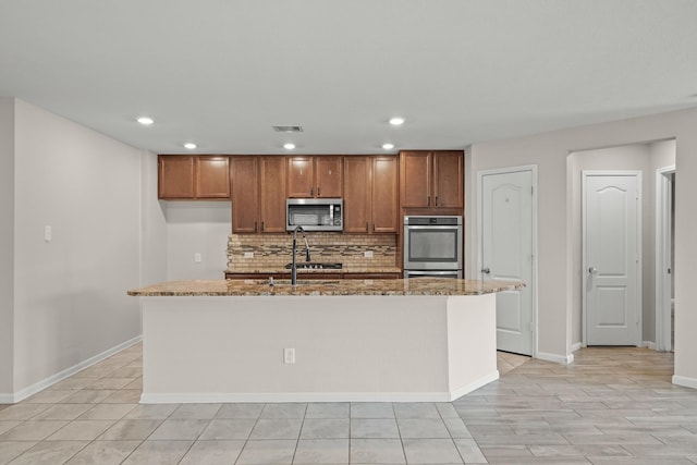 kitchen with appliances with stainless steel finishes, light stone countertops, an island with sink, and decorative backsplash