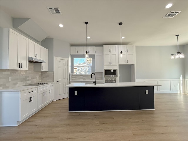 kitchen with an island with sink, stainless steel microwave, decorative light fixtures, white cabinets, and sink