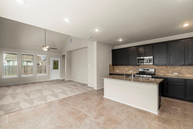 kitchen featuring vaulted ceiling, decorative backsplash, an island with sink, light stone counters, and stainless steel appliances