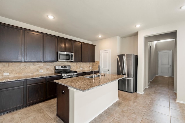 kitchen with light stone countertops, appliances with stainless steel finishes, dark brown cabinets, a kitchen island with sink, and sink