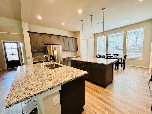 kitchen with light stone countertops, sink, stainless steel refrigerator with ice dispenser, an island with sink, and pendant lighting