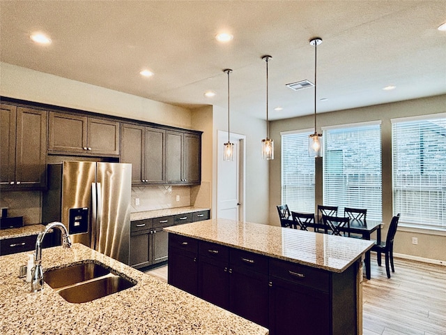 kitchen featuring pendant lighting, sink, stainless steel refrigerator with ice dispenser, an island with sink, and light stone counters