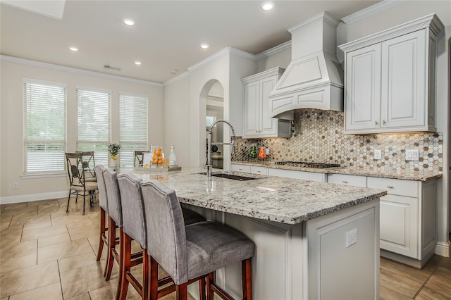 kitchen with light stone counters, custom exhaust hood, sink, stainless steel gas stovetop, and an island with sink