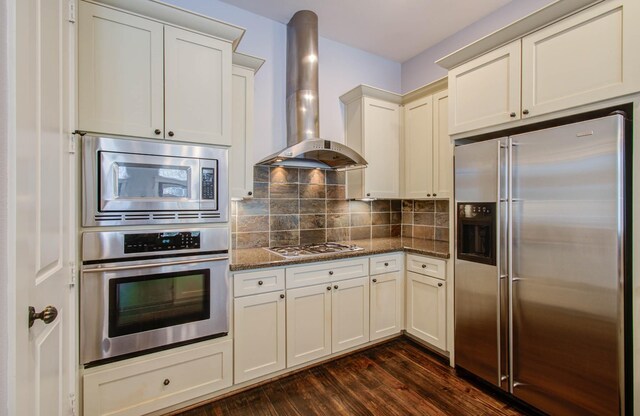 kitchen featuring dark wood-type flooring, dark stone counters, wall chimney exhaust hood, tasteful backsplash, and stainless steel appliances