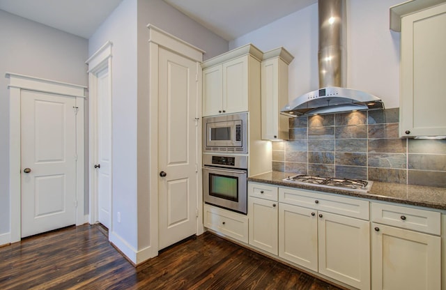 kitchen with appliances with stainless steel finishes, dark hardwood / wood-style flooring, backsplash, wall chimney exhaust hood, and dark stone countertops