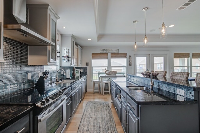 kitchen with a tray ceiling, sink, wall chimney range hood, stainless steel electric range oven, and an island with sink