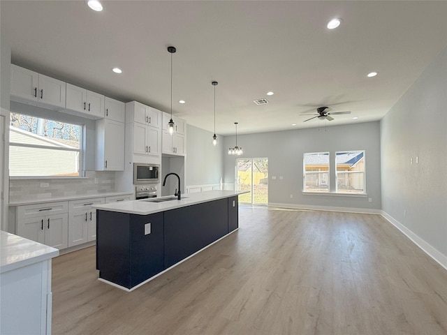 kitchen featuring an island with sink, stainless steel appliances, pendant lighting, ceiling fan with notable chandelier, and white cabinets