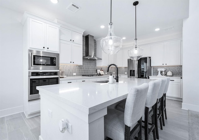 kitchen featuring wall chimney range hood, hanging light fixtures, an island with sink, white cabinetry, and stainless steel appliances