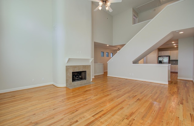 unfurnished living room with a tile fireplace, ceiling fan, a towering ceiling, and light wood-type flooring