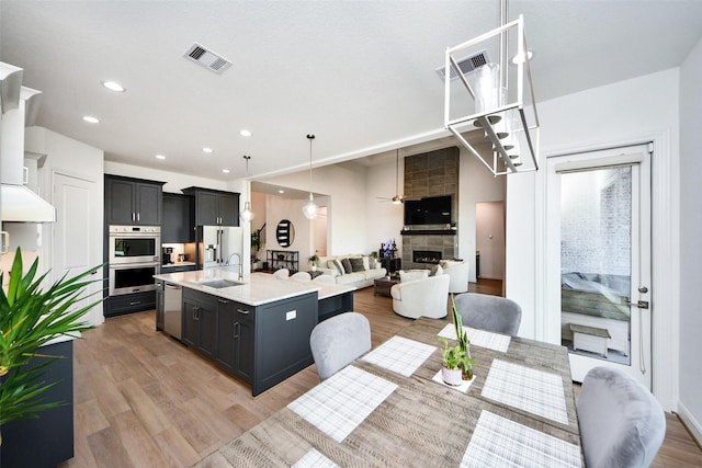 kitchen featuring appliances with stainless steel finishes, a large fireplace, a center island with sink, light hardwood / wood-style flooring, and hanging light fixtures