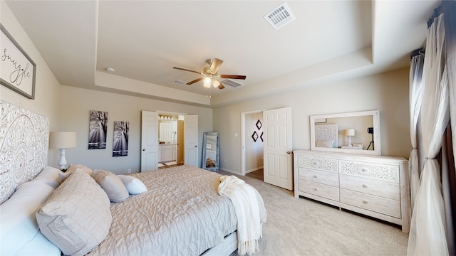 bedroom with ceiling fan, a raised ceiling, light colored carpet, and ensuite bathroom