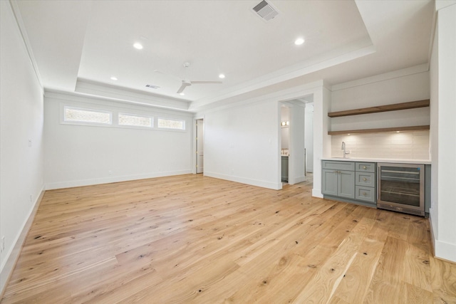 unfurnished living room with sink, ceiling fan, wine cooler, a tray ceiling, and light hardwood / wood-style floors