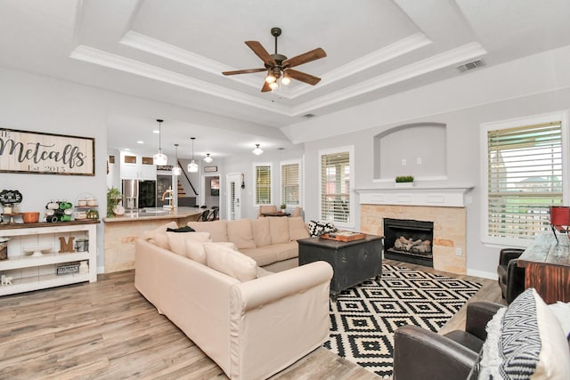 living room featuring a tray ceiling, a stone fireplace, ceiling fan, and light hardwood / wood-style flooring