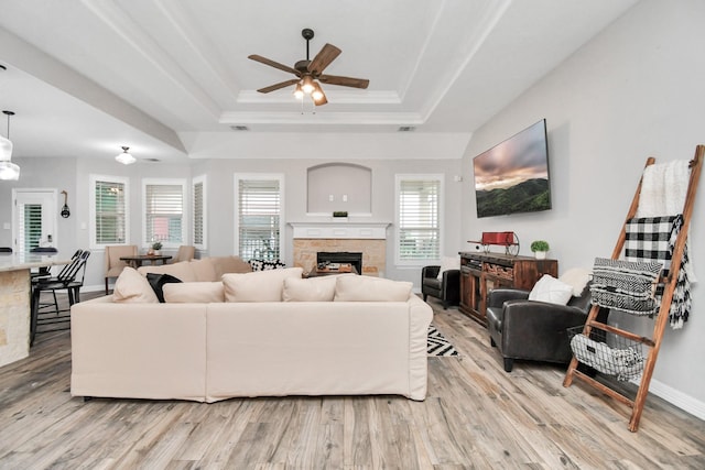 living room featuring ceiling fan, a stone fireplace, a raised ceiling, and light wood-type flooring