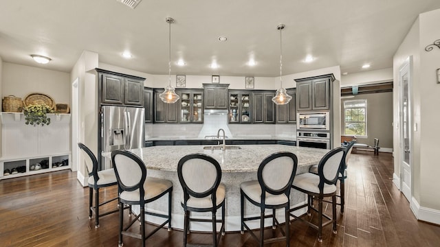 kitchen with sink, pendant lighting, stainless steel appliances, light stone countertops, and a kitchen island with sink