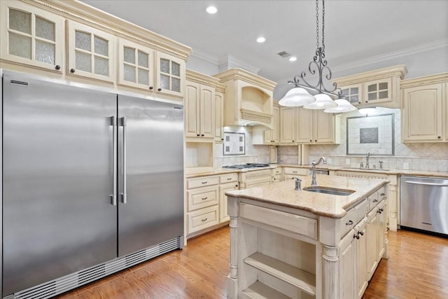 kitchen with stainless steel appliances, sink, pendant lighting, cream cabinetry, and an island with sink