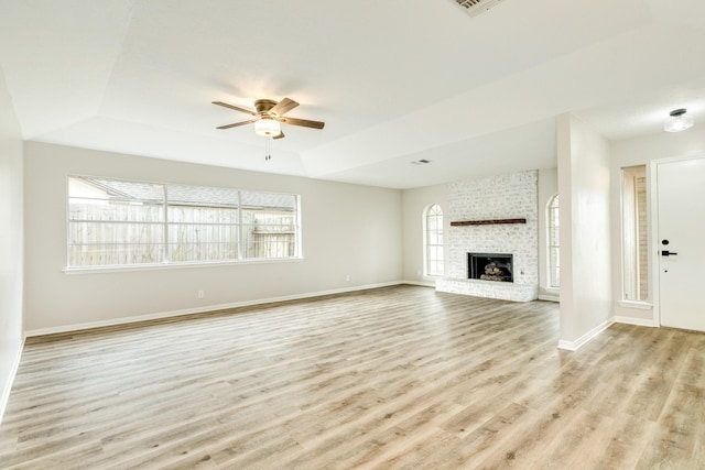 unfurnished living room with a brick fireplace, a tray ceiling, ceiling fan, and light wood-type flooring