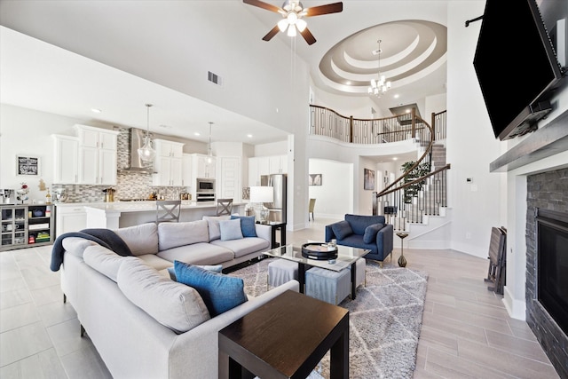 living room with ceiling fan with notable chandelier, a stone fireplace, a high ceiling, and a tray ceiling