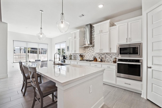 kitchen featuring a center island with sink, sink, wall chimney exhaust hood, appliances with stainless steel finishes, and decorative light fixtures