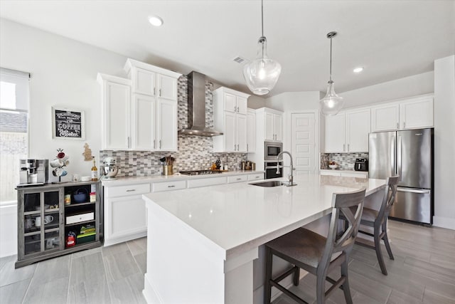 kitchen with stainless steel appliances, sink, wall chimney range hood, decorative light fixtures, and a center island with sink