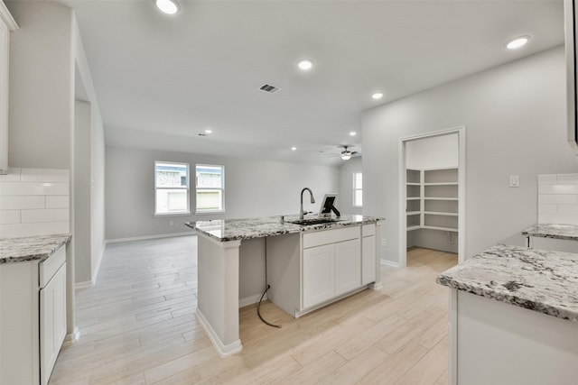 kitchen with white cabinets, a center island with sink, sink, ceiling fan, and light stone countertops