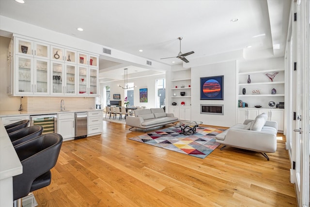 living room with wet bar, ceiling fan with notable chandelier, wine cooler, built in features, and light hardwood / wood-style floors