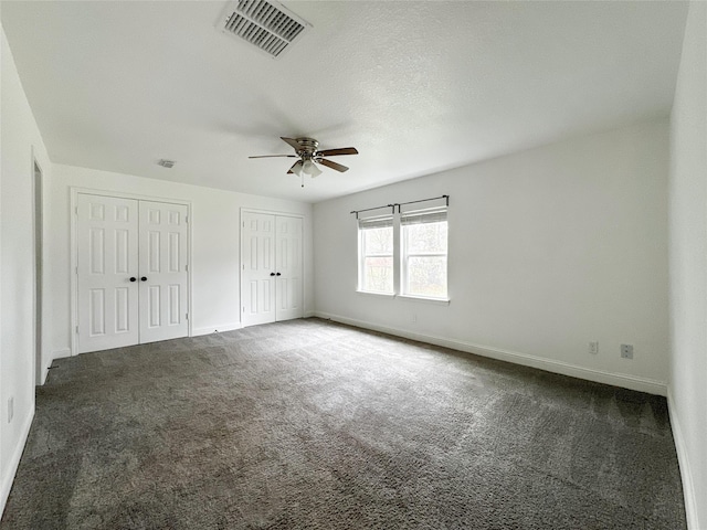 unfurnished bedroom with ceiling fan, two closets, a textured ceiling, and dark carpet