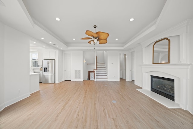 unfurnished living room with a glass covered fireplace, stairway, light wood-style floors, crown molding, and a raised ceiling