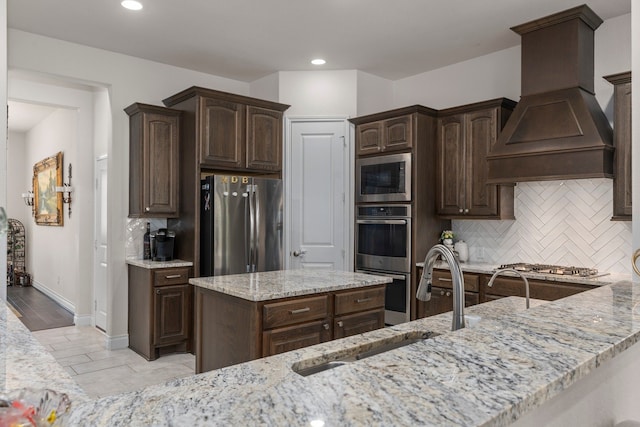 kitchen with dark brown cabinetry, light stone counters, decorative backsplash, custom range hood, and appliances with stainless steel finishes