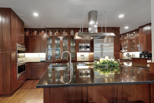 kitchen with sink, built in appliances, dark stone countertops, pendant lighting, and island range hood