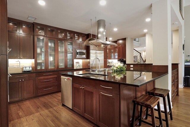 kitchen with a center island with sink, sink, built in appliances, island range hood, and wood-type flooring